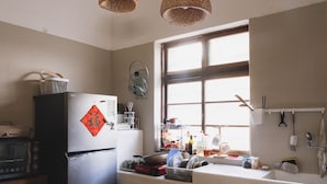 A kitchen with a refrigerator, microwave, and various kitchen utensils. There is a window providing natural light and a hanging light fixture. - TUNGWEI38 HOSTEL (Magong)