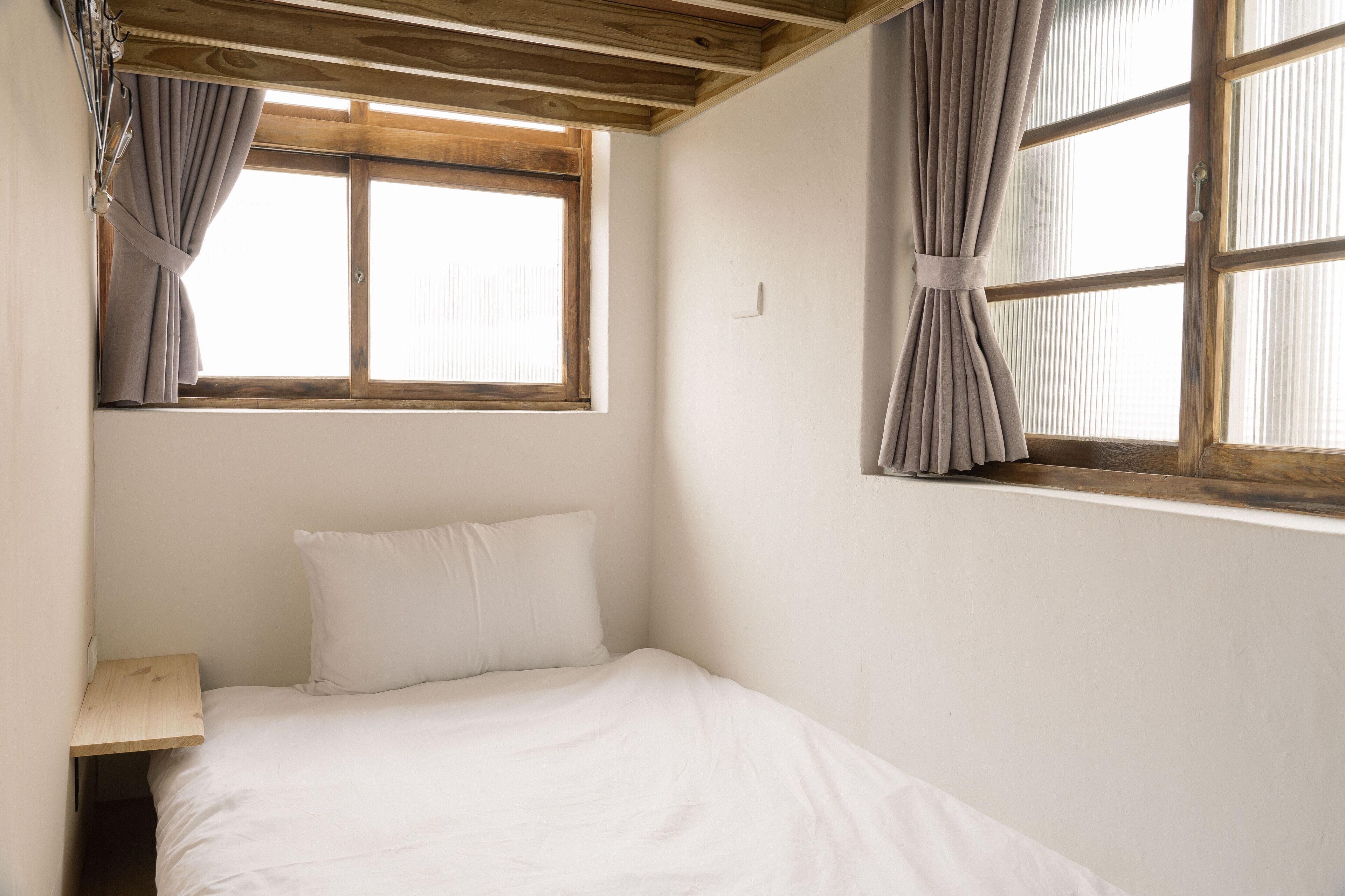 Ein kleines Zimmer mit einem Einzelbett, Holzboden, einem Fenster mit Vorhängen und einem Nachttisch.