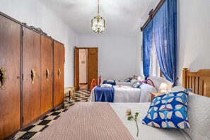 Une chambre avec un sol à carreaux, une armoire en bois, deux lits avec une literie bleue et blanche, un lustre et une fenêtre avec des rideaux bleus.
