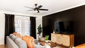A modern living room with a grey sofa, black curtains, a ceiling fan, a TV on a wooden cabinet, and a plant.