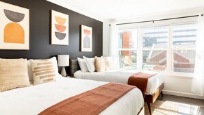 A hotel room with two beds, a large window with a view of buildings, and artwork on the wall.