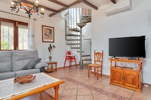 A living room with a sofa, a coffee table, a TV on a wooden stand, a red chair, a spiral staircase, and a painting on the wall. - Holiday Home "Torre del Manzano" with Mountain View, Private Pool & Wi-Fi (Torrox)
