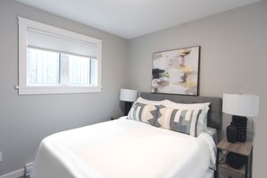 Quarto com cama, duas luminárias de cabeceira, janela com persianas e um quadro abstrato pendurado na parede.