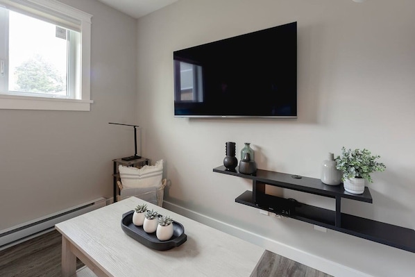 Sala de estar com televisão de tela plana na parede, mesa de madeira com objetos decorativos e uma cadeira.