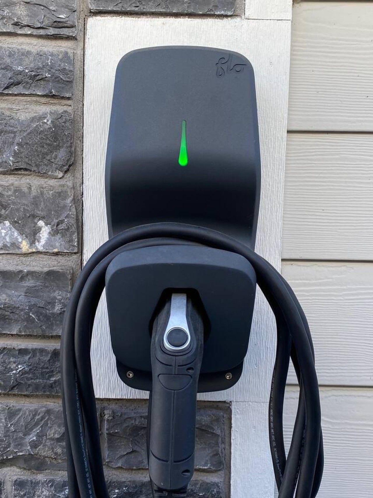 A black electric vehicle charging station with a green light indicator, mounted on a stone wall.