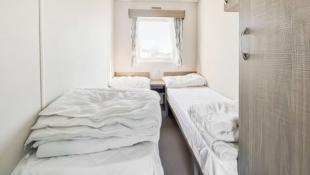 Une petite chambre propre, avec deux lits simples, une fenêtre avec des rideaux et une porte.