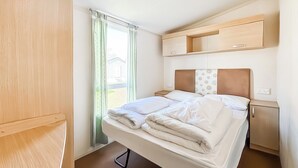 Une pièce de taille modeste comprenant un lit, une petite table de chevet, une étagère intégrée et une fenêtre avec des rideaux.