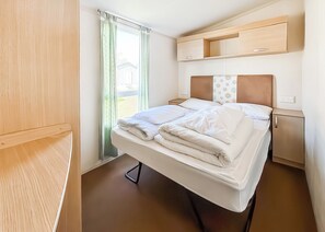 A compact room with a bed, a small bedside table, a built-in shelf, and a window with curtains.