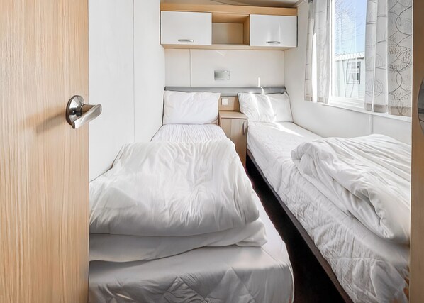 A compact bedroom with two beds, a small bedside table, a cabinet, and a window with curtains.