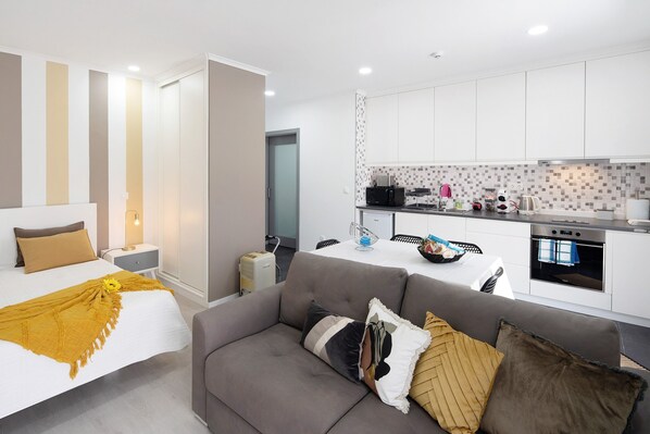 A modern hotel room with a bed, a sofa, a dining area, and a kitchenette.
