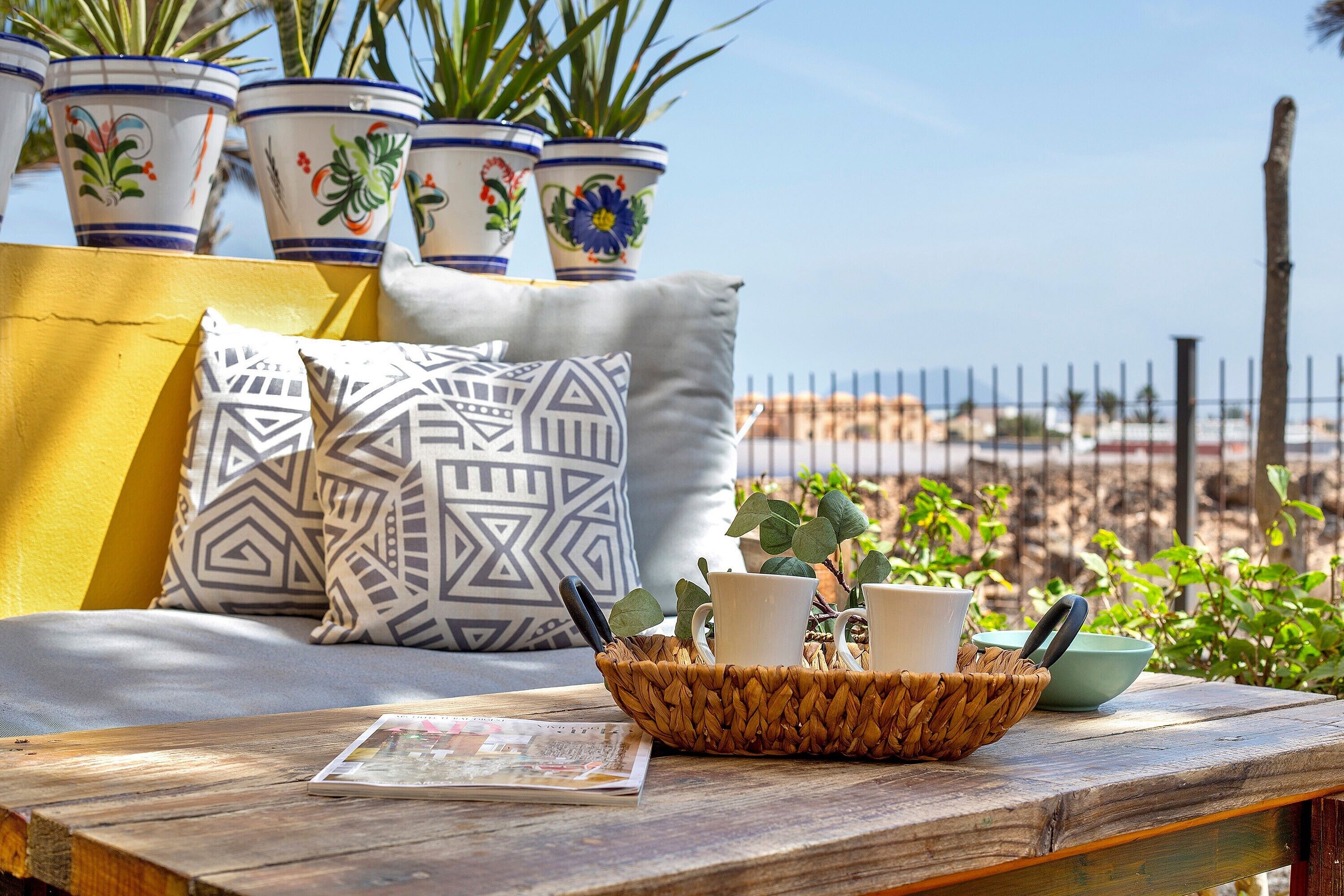 A wicker basket with cups and a bowl on a wooden table, a sofa with patterned cushions, and potted plants in the background.