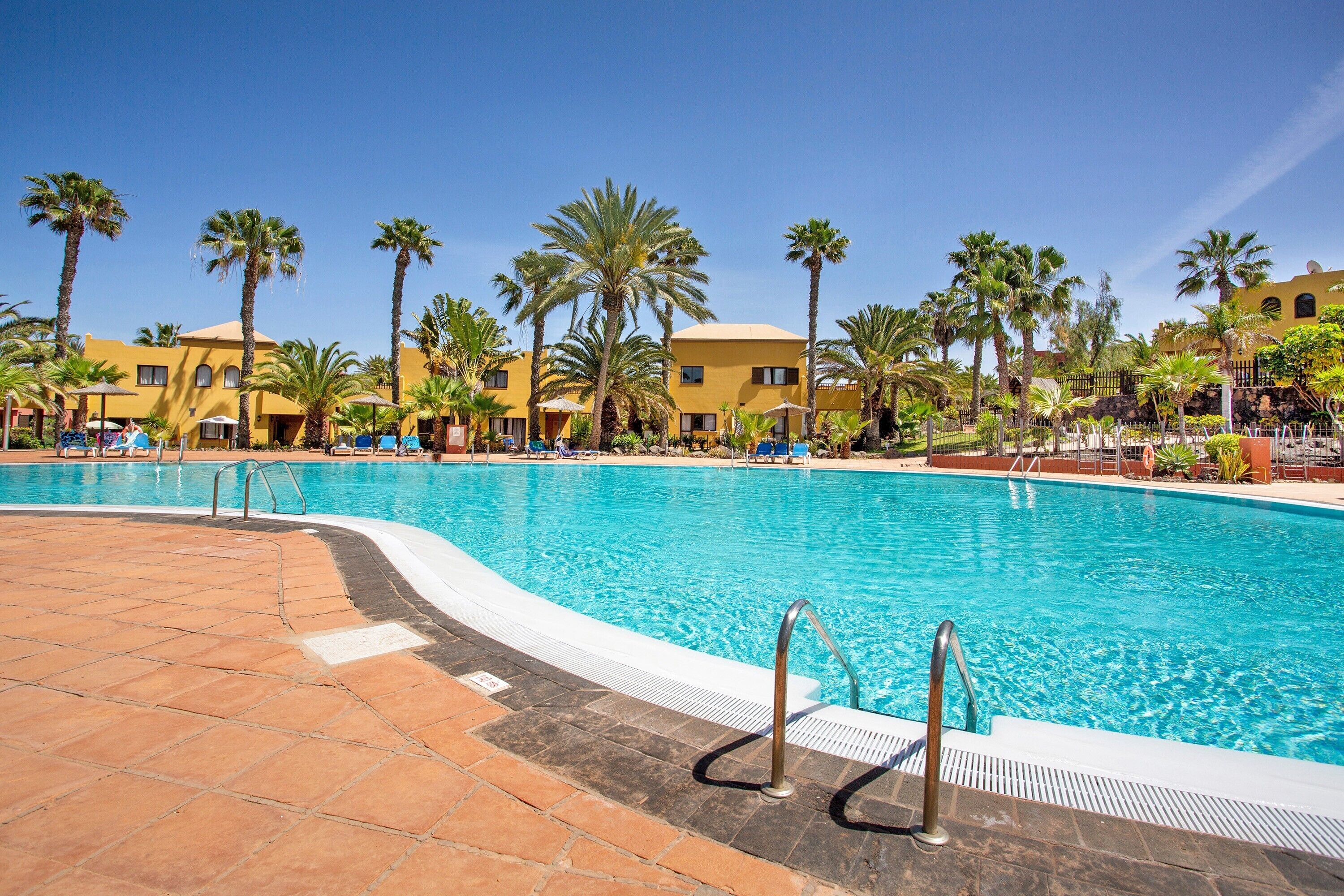 A swimming pool with a tiled deck, palm trees, and yellow buildings in the background.