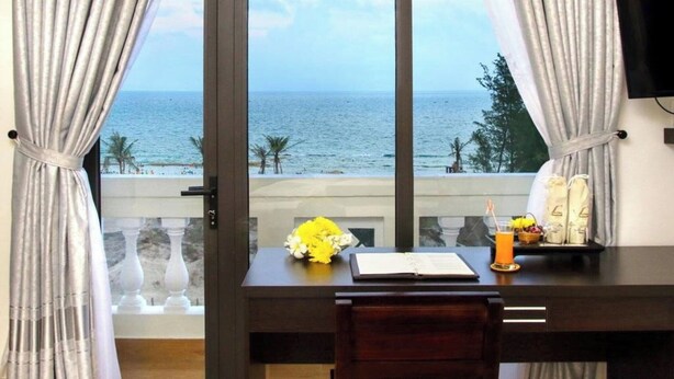 Una stanza con vista sul mare, una scrivania con un libro, un bicchiere di succo d'arancia e un bouquet di fiori.