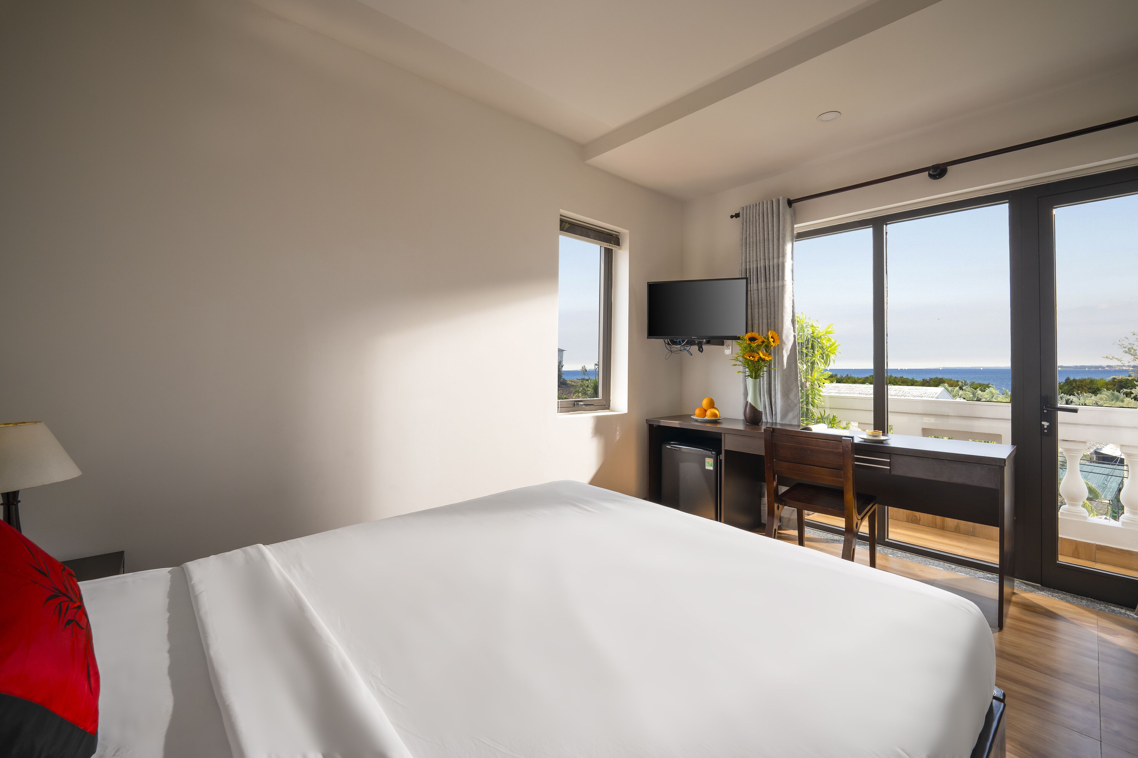 A hotel room with a bed, a desk, a television, and a large window offering a view of the sea.