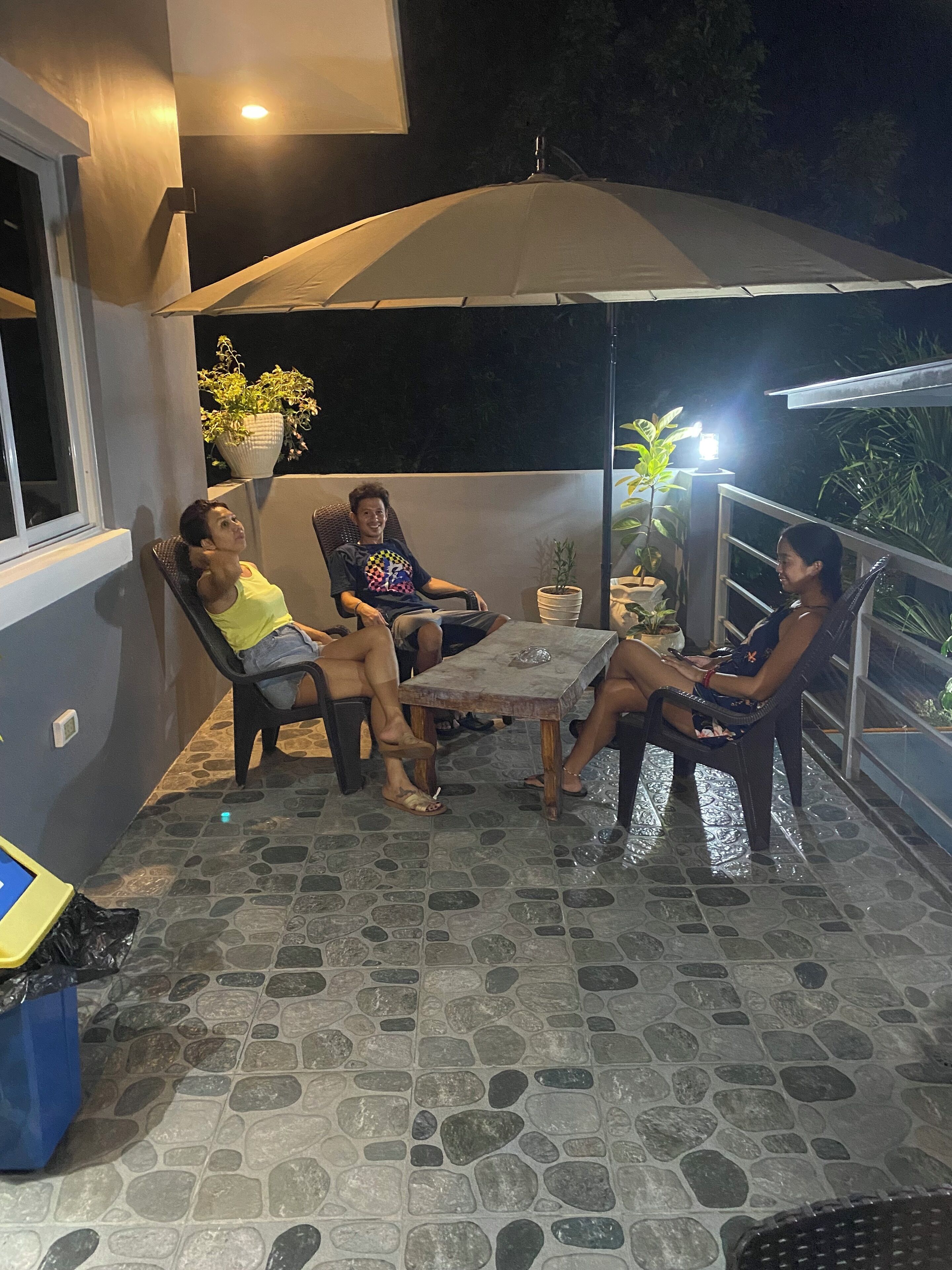 A group of people seated around a table on a patio with a tiled floor and an umbrella providing shade.