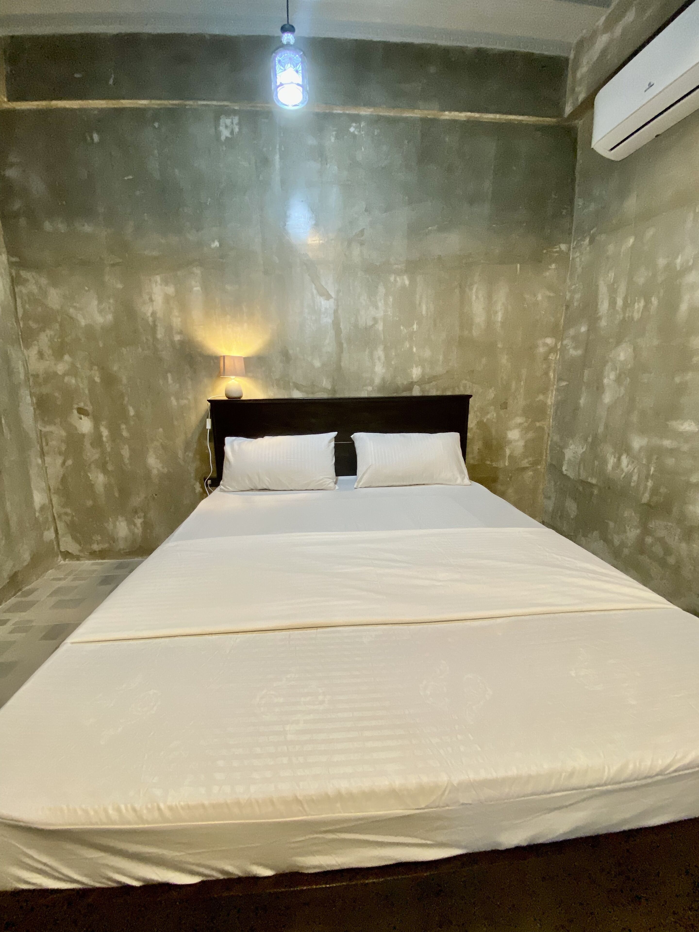 Una cama individual con sábanas blancas en una habitación con pared de concreto y un equipo de aire acondicionado.