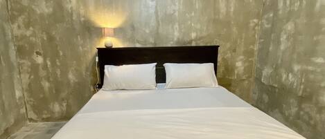 Una cama individual con sábanas blancas en una habitación con pared de concreto y un equipo de aire acondicionado.