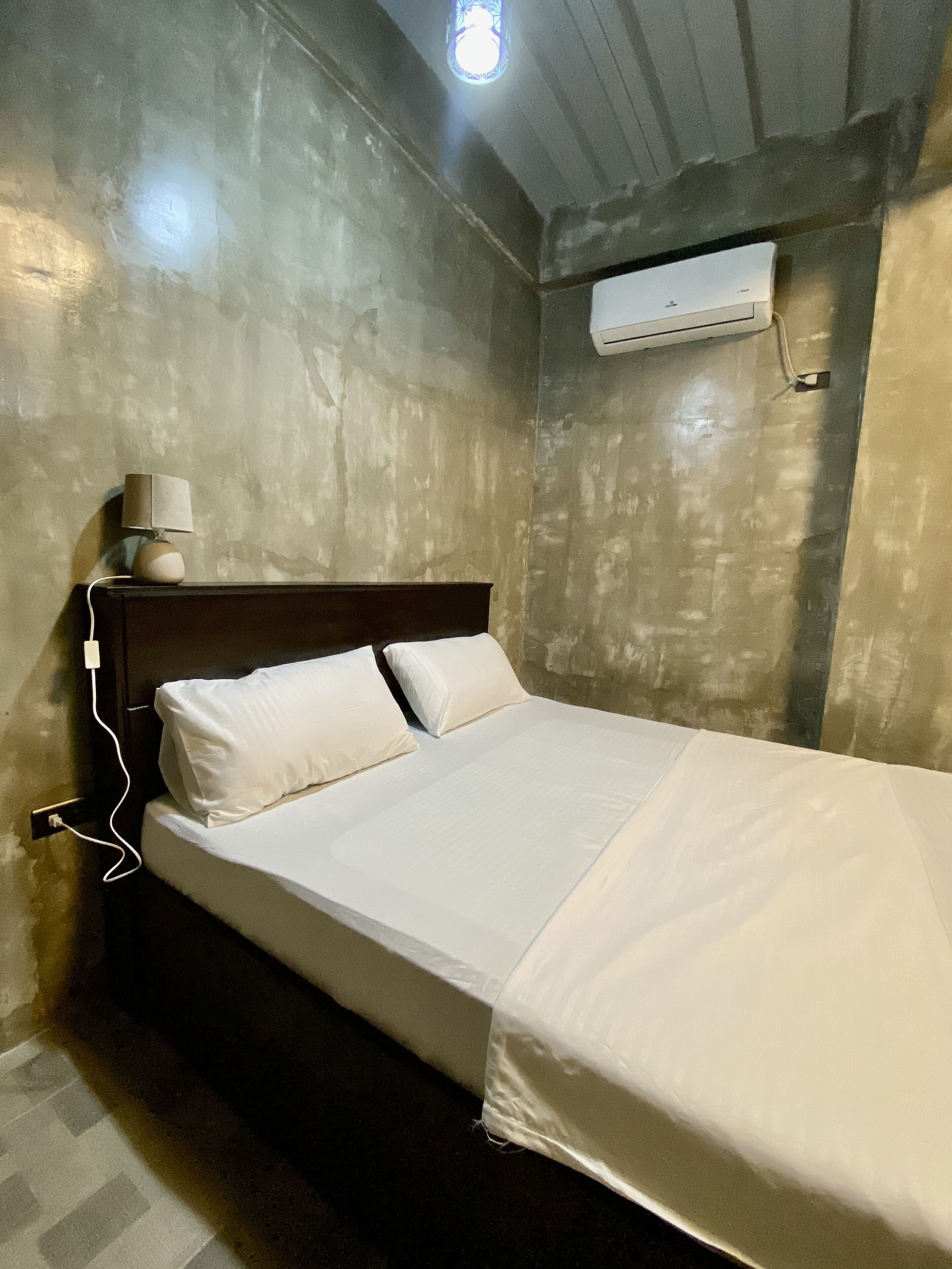 A single bed with white linens, a bedside lamp, and an air conditioning unit mounted on the wall.