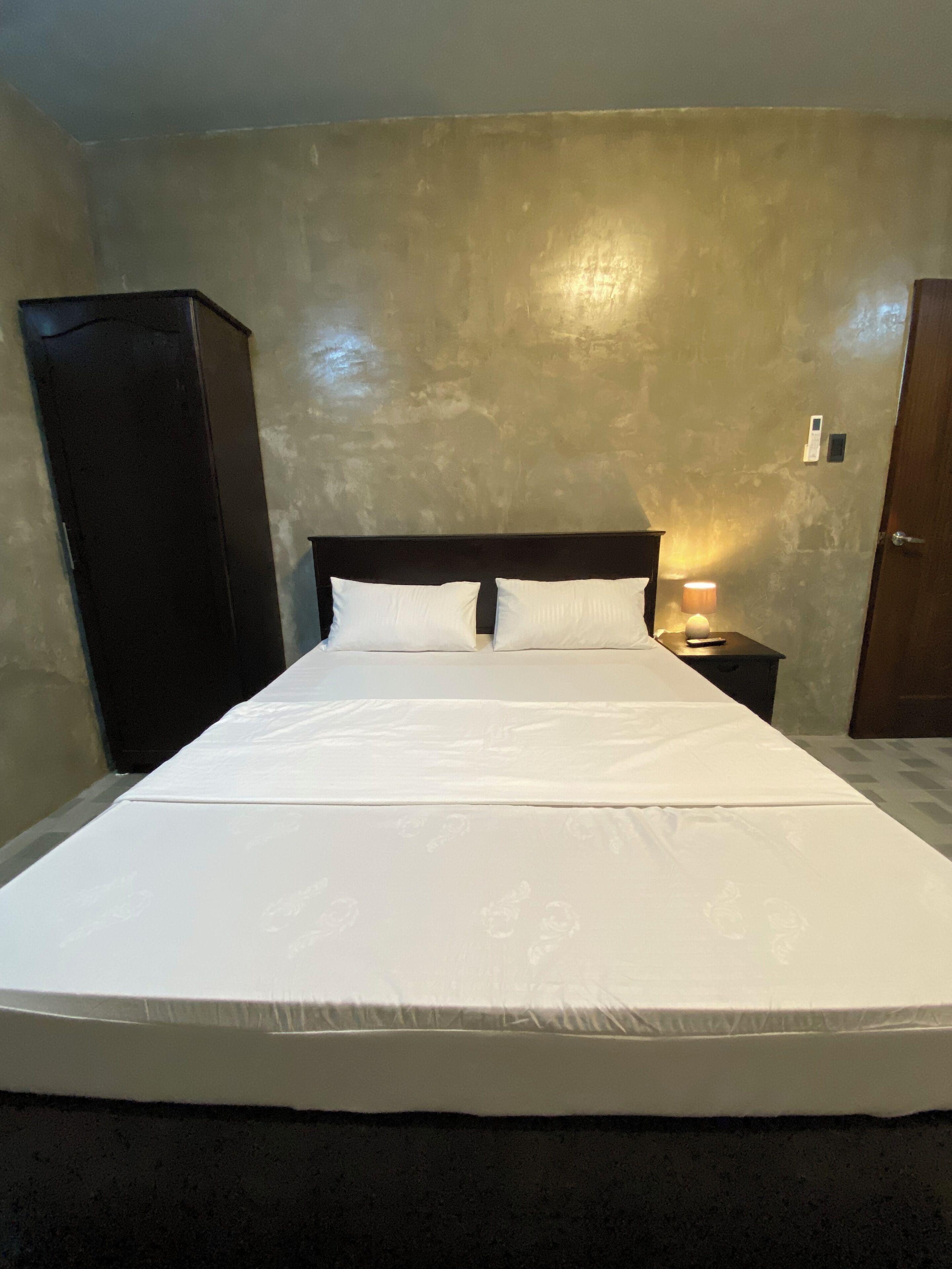 A single bed with white bedding in a room with a textured beige wall, a wooden wardrobe, and a nightstand with a lamp.