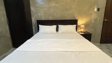 A single bed with white bedding in a room with a textured beige wall, a wooden wardrobe, and a nightstand with a lamp.