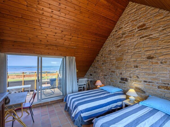 A bedroom with two beds, a wooden ceiling, a stone wall, a balcony with a view of the sea, and a table with chairs.
