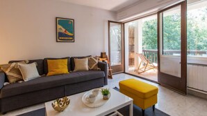 A modern living room with a sofa, a coffee table, and a yellow ottoman. There is a large window with a view of greenery outside. - Le Bourret by Interhome (Hossegor)