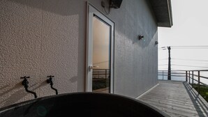 A building with a textured exterior wall, a door leading to an interior space, and a wooden deck with a railing overlooking a body of water. - SUNNY SIDE HOUSE (Ishikari)