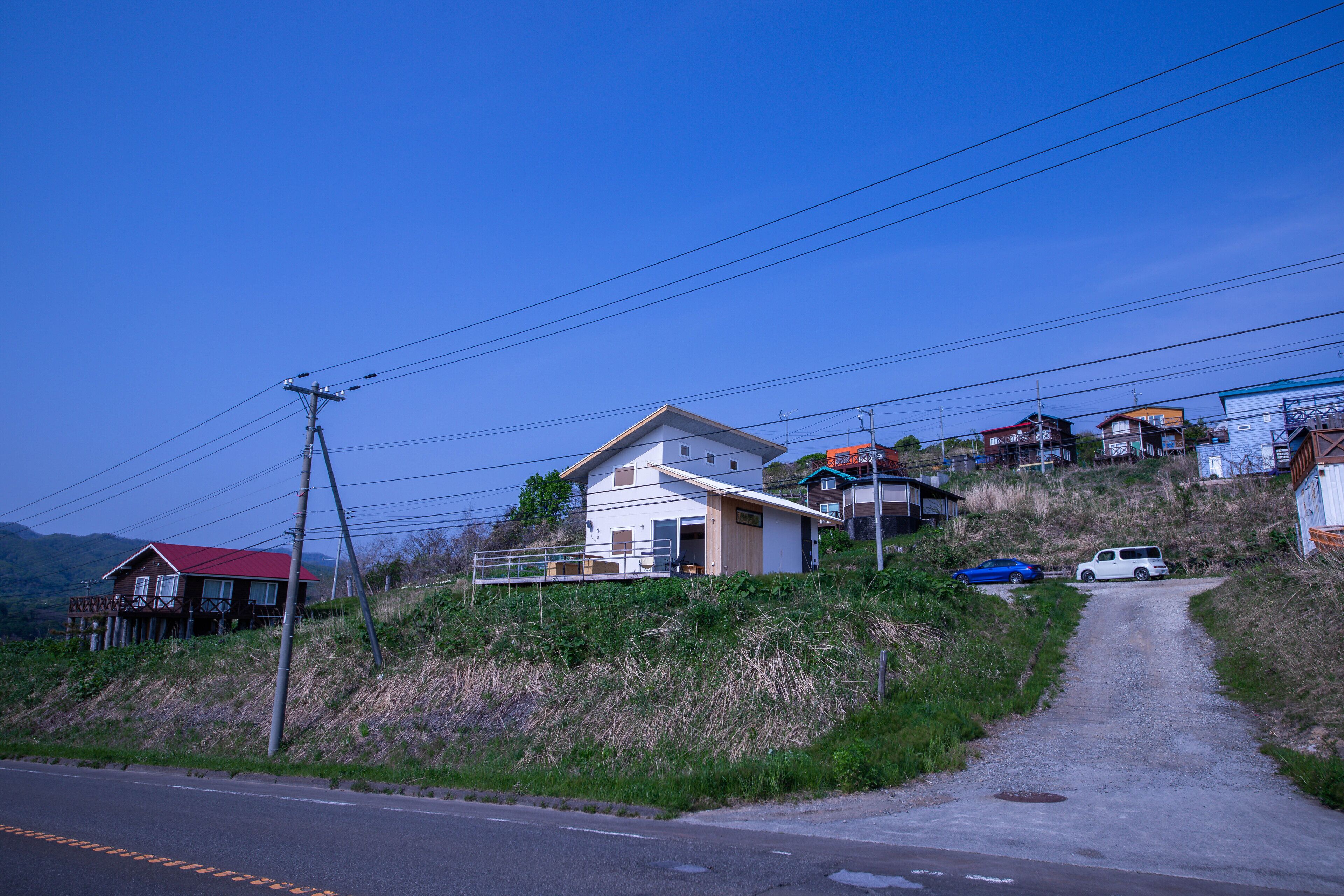A rural road leading to a house with a red roof, surrounded by hills and other houses.