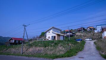 A rural road leading to a house with a red roof, surrounded by hills and other houses.