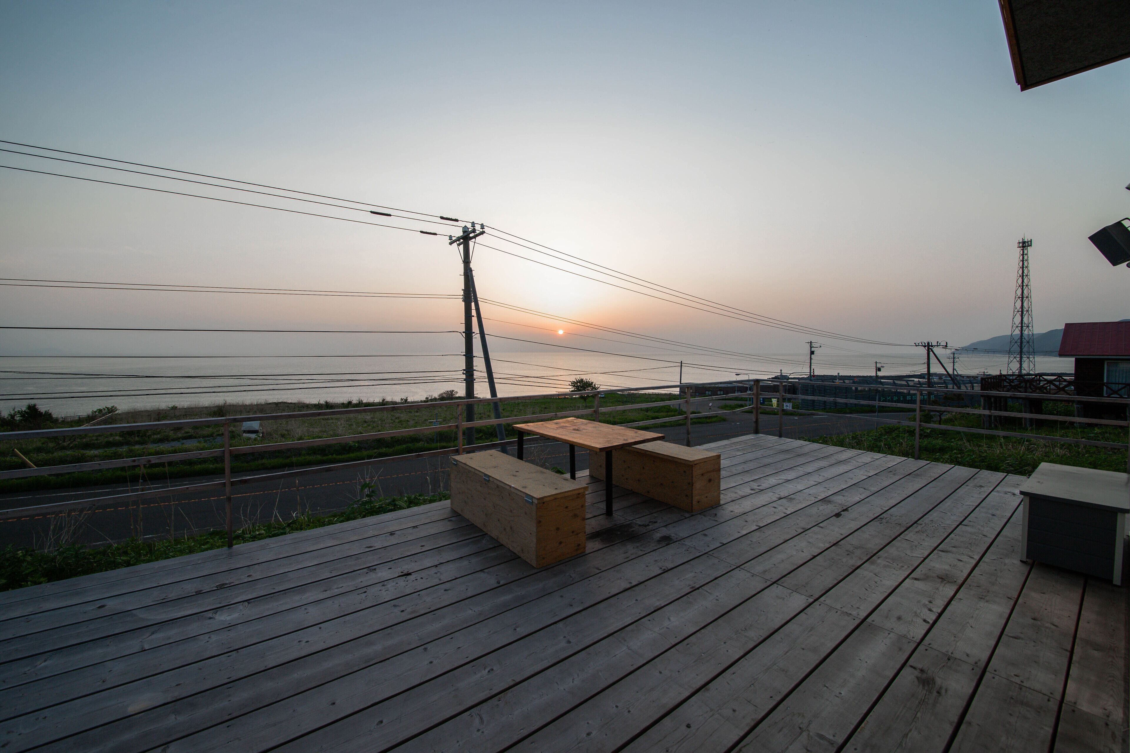 A wooden deck with a table and benches, overlooking a road and power lines, with a sunset in the background.