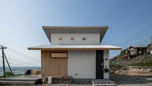 A modern single-story building with a flat roof, a wooden deck with seating, and a view of the sea. - SUNNY SIDE HOUSE (Ishikari)