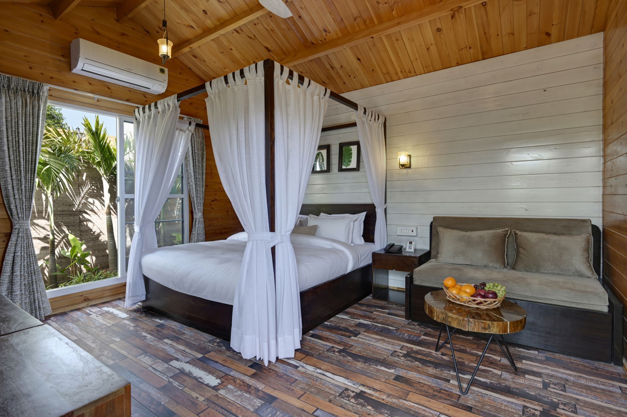 Quarto com teto de madeira, cama com roupa de cama branca, piso de madeira, sofá, mesa de cabeceira e uma cesta de frutas.