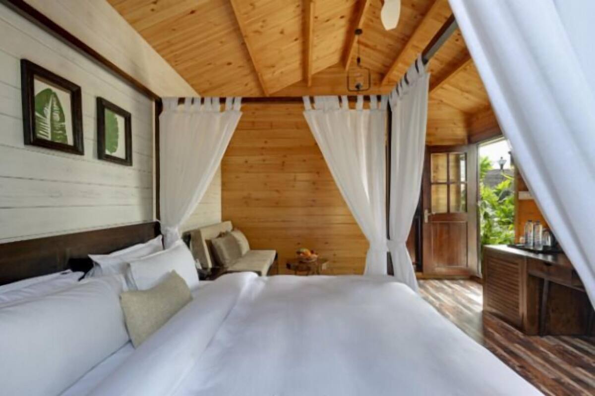 Een slaapkamer in een houten hutje, met een bed, gordijnen en een klein tafeltje met een lamp.