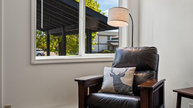 Una habitación con ventana, una silla con un cojín de ciervo y una lámpara.
