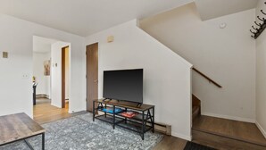 A modern living room with a flat-screen TV on a wooden stand, a patterned rug, and a staircase.