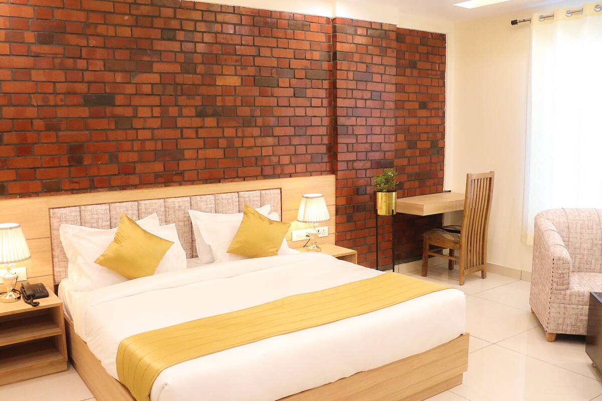 A hotel room with a brick-patterned headboard, a large bed with white and yellow bedding, a desk with a chair, and a small plant.