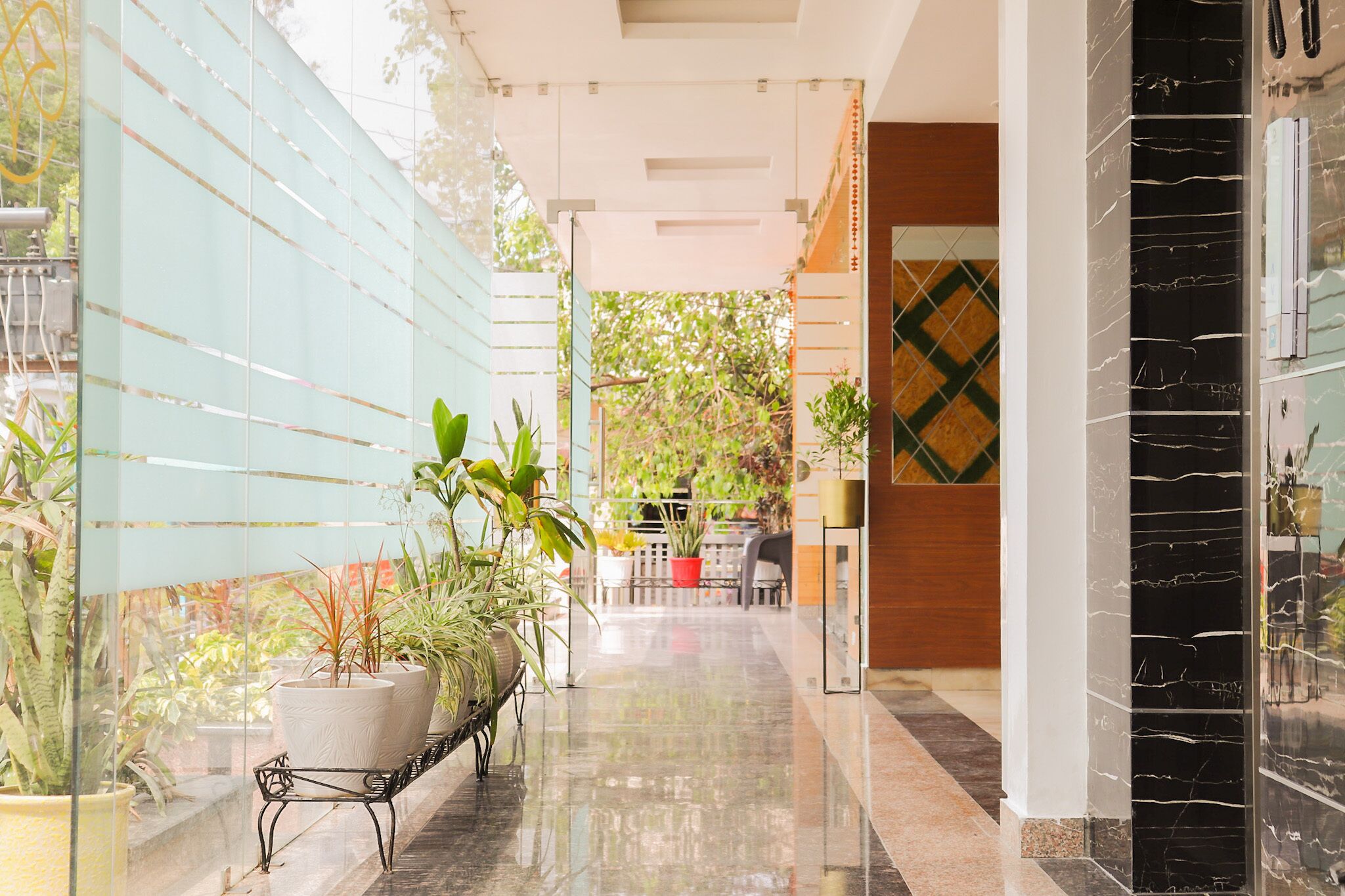 A corridor with potted plants, a bench, and a view of an outdoor area with trees and a table.
