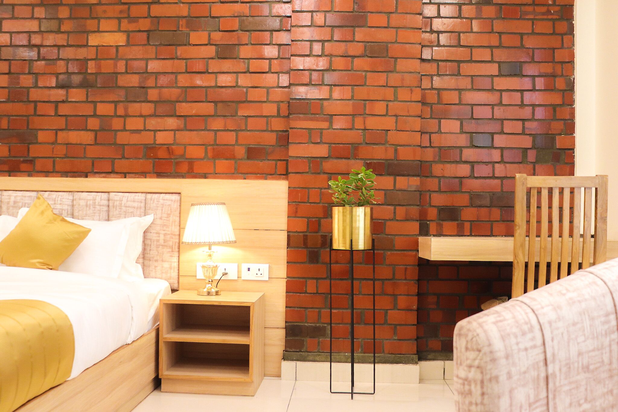 Ein Hotelzimmer mit Backsteinwand, einem Bett mit weißer Bettwäsche und einer gelben Decke, einem Nachttisch mit Lampe, einer kleinen Pflanze und einem Holzstuhl.