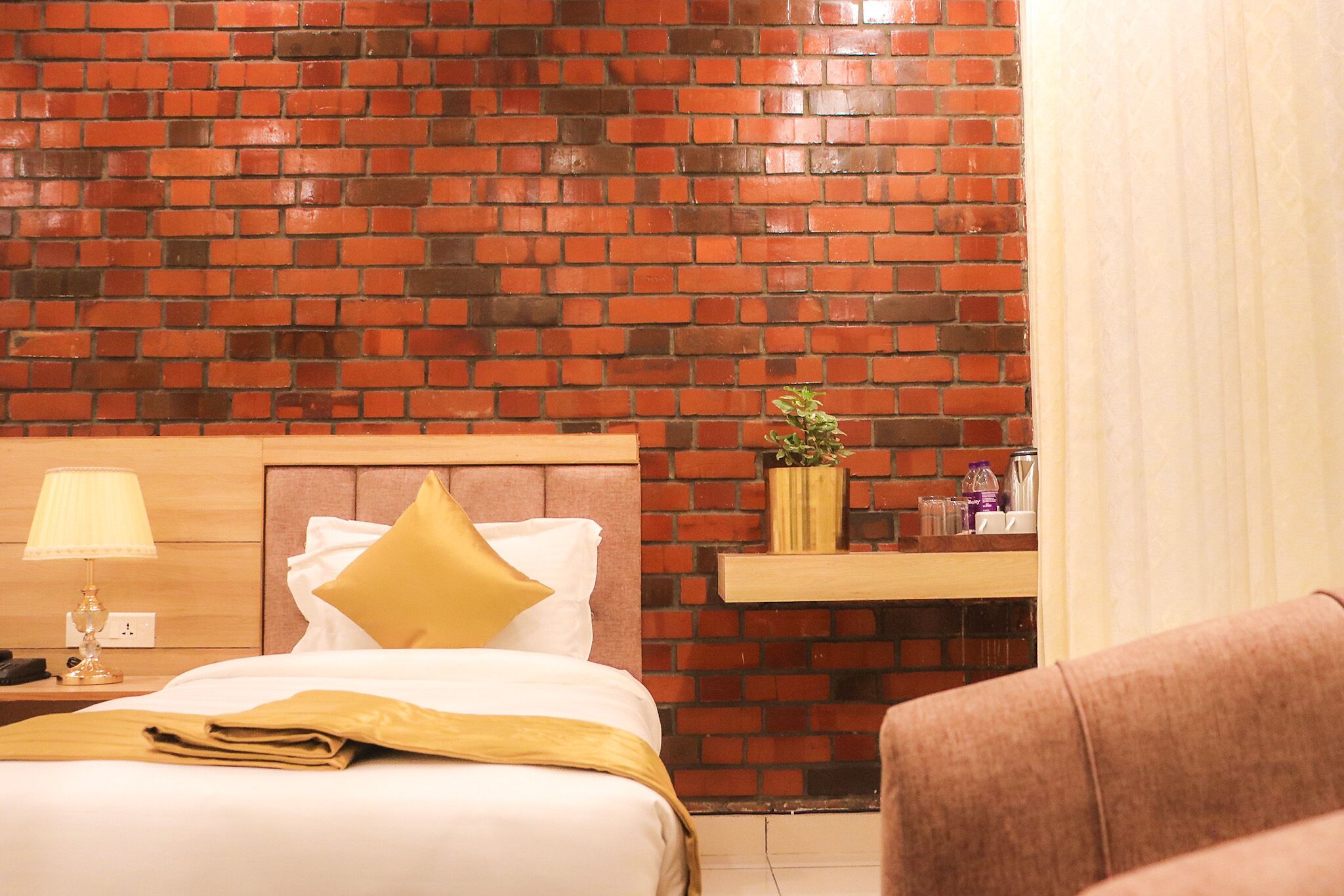 A hotel room with a brick-patterned wall, a bed with white linens and a yellow pillow, a bedside table with a lamp, and a small shelf with a plant and toiletries.