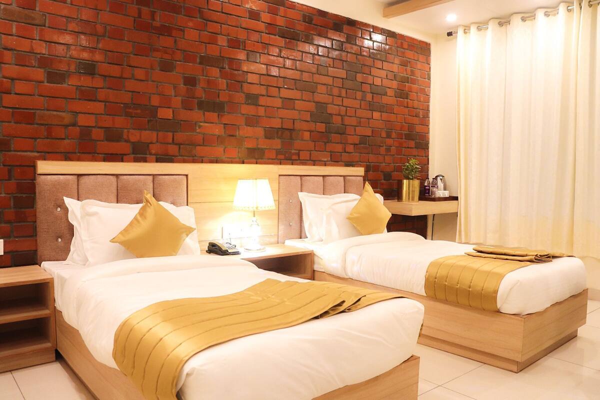 A hotel room with two beds, a brick wall, a nightstand with a lamp, and a window with curtains.