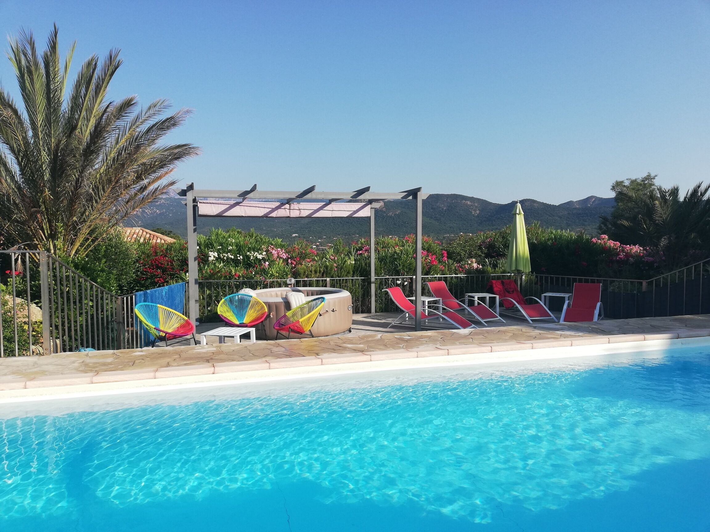 En pool med kristallklart blått vatten, omgiven av röda solstolar och färgglada stolar, med en pergola och utsikt över berg i bakgrunden.