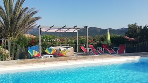 A swimming pool with clear blue water, surrounded by red lounge chairs and colorful chairs, with a pergola and a view of mountains in the background. - Appartement Dans Villa Piscine ET SPA (SAINT LUCIE DE PORTO VECCHIO)