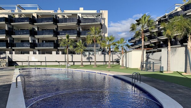 Una piscina de borde curvo, rodeada por un muro bajo y un área verde. Al fondo, edificios de varios pisos con balcones y palmeras.