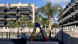 Un parque infantil con columpios y tobogán, rodeado de palmeras y edificios modernos.