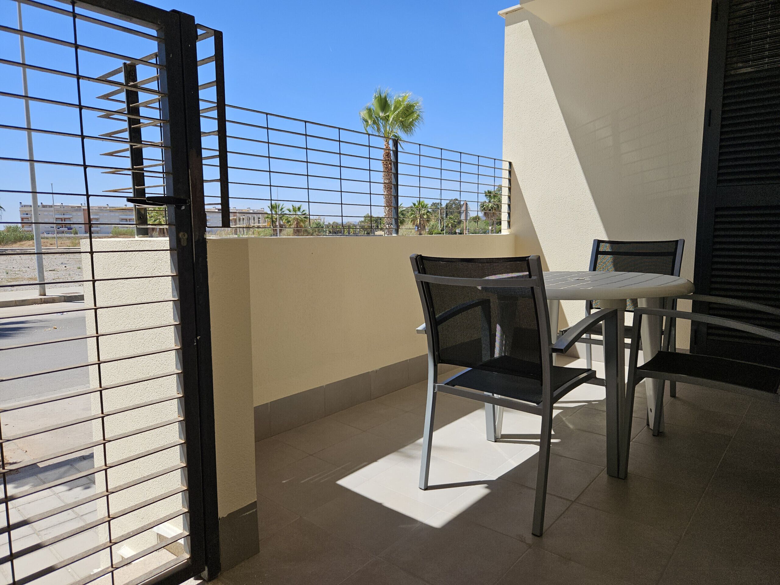 A balcony with a metal gate, a table, two chairs, and a view of a palm tree and buildings in the background.