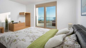 Quarto moderno com uma cama grande, uma mesa de cabeceira de madeira, uma pequena mesa e vista para o mar através de uma porta de correr de vidro.