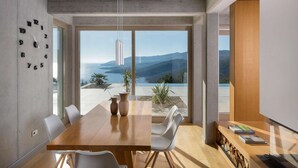 Uma área de refeições moderna com uma mesa de madeira, cadeiras brancas e vista para o mar e montanhas através de grandes janelas.