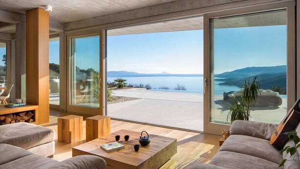 Sala moderna com uma ampla porta de vidro deslizante que dá para uma vista panorâmica de um lago e montanhas. Há uma mesa de centro de madeira com livros e uma chaleira, e um sofá com uma guitarra encostada.