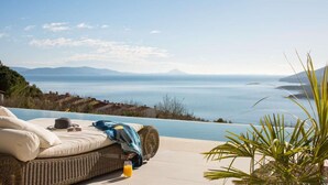 Uma cadeira de vime com almofada e chapéu, um copo de suco de laranja, e vista para o mar e montanhas ao longe.