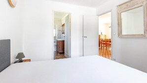 Un dormitorio con cama, una lámpara de noche, una puerta que da a un baño y un comedor visible a través de una puerta abierta.